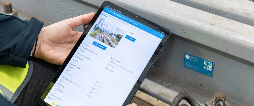 Ein Instandhaltungsmitarbeiter hält ein Tablet zu einem am Bahngleis angebrachten Sensor